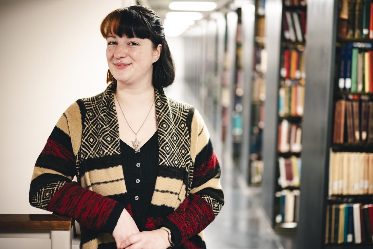 Olivia Mathers stands in the stacks and poses for a photo. She wears a stripped cardigan.